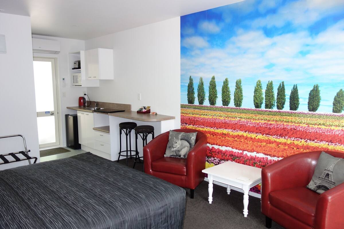 A modern room with a bed, two red leather chairs, a small white table, a kitchen area, and a large floral mural on the wall.