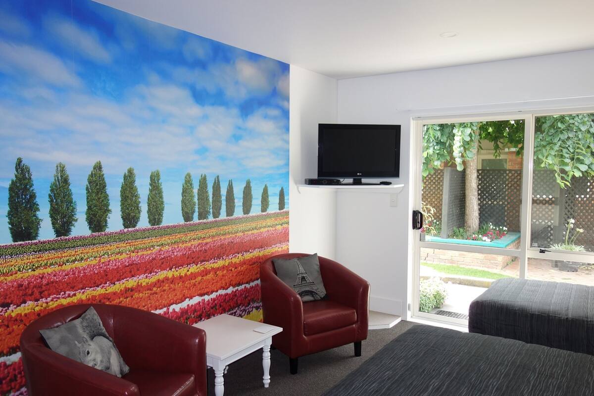 A room with a large window, a flat-screen TV, a red armchair, a white table, and a colorful mural on the wall.