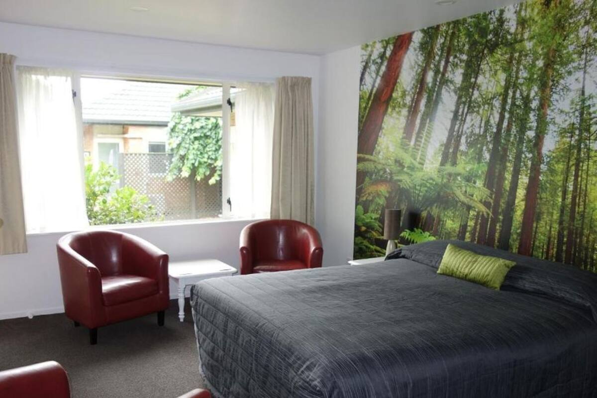 A hotel room with a bed, two red chairs, a small table, and a large window with curtains.