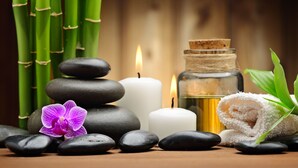 A stack of smooth stones, a lit candle, a bottle of oil, and a rolled towel, suggesting a spa setting.