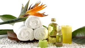 A spa setting with a rolled white towel, a green candle, a bottle of oil, and a yellow candle on a bed of white pebbles.