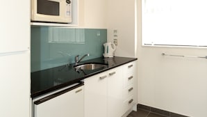 A compact kitchen with white cabinets, a black countertop, a microwave, a kettle, and a window with curtains. - Gothic Heights Motel (Christchurch)