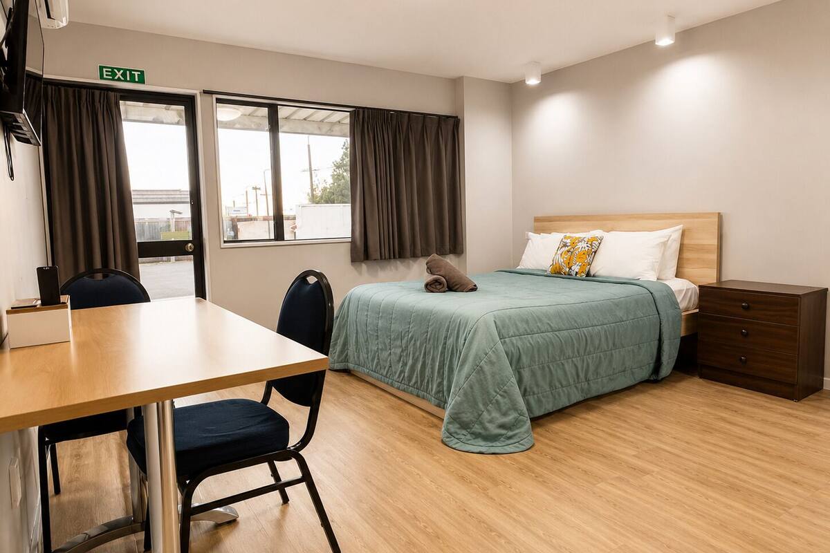 A hotel room with a bed, bedside table, chair, desk, and an exit sign.