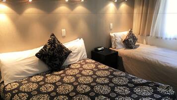 Habitación de hotel con dos camas, cada una con ropa de cama y almohadas estampadas, una mesita de noche con control remoto e iluminación empotrada en la pared.