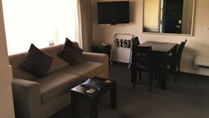 Un salon comprenant un canapé, une table basse, un téléviseur fixé au mur, une table à manger avec des chaises et un grand miroir.