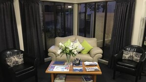 Un salon avec un canapé beige, deux fauteuils en cuir noir, une table basse avec des magazines et un vase de fleurs, ainsi que de grandes fenêtres avec des rideaux.