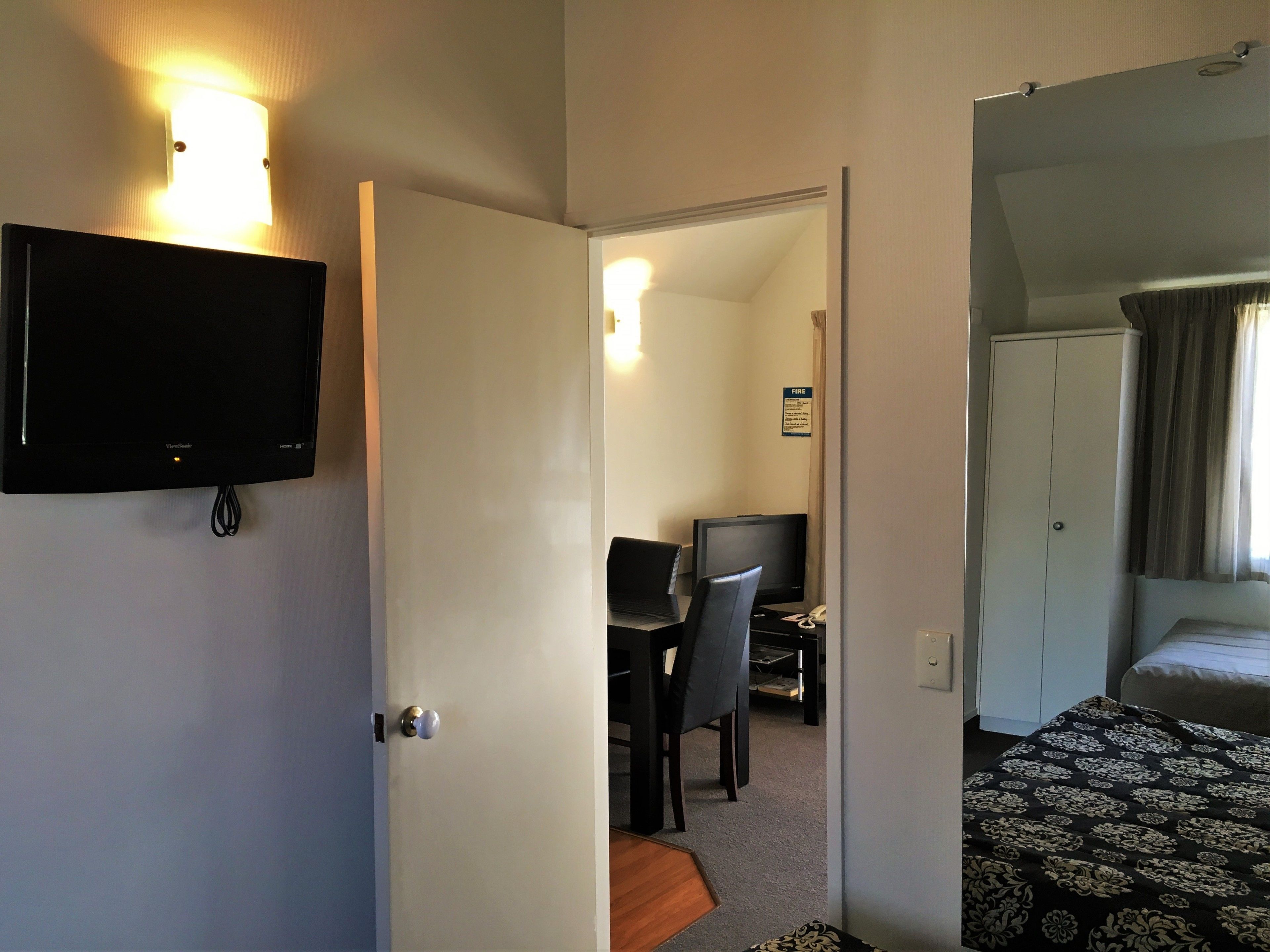 Una habitación de hotel con una cama, un escritorio con computadora, un televisor en la pared y una puerta que da a otra habitación.
