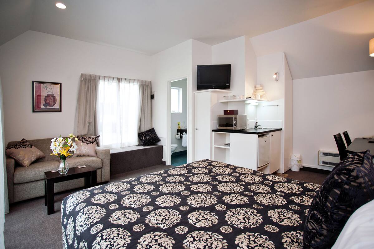 Ein Hotelzimmer mit Bett, einer Couch, einem kleinen Tisch mit Blumenvase, einer Küchenzeile und einem an der Wand befestigten Fernseher.