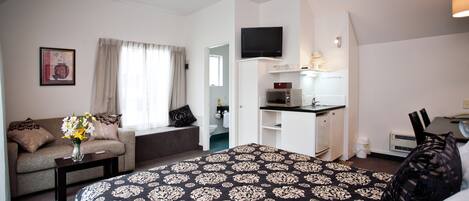 A hotel room with a bed, a sofa, a small table with a vase of flowers, a kitchenette, and a TV mounted on the wall.