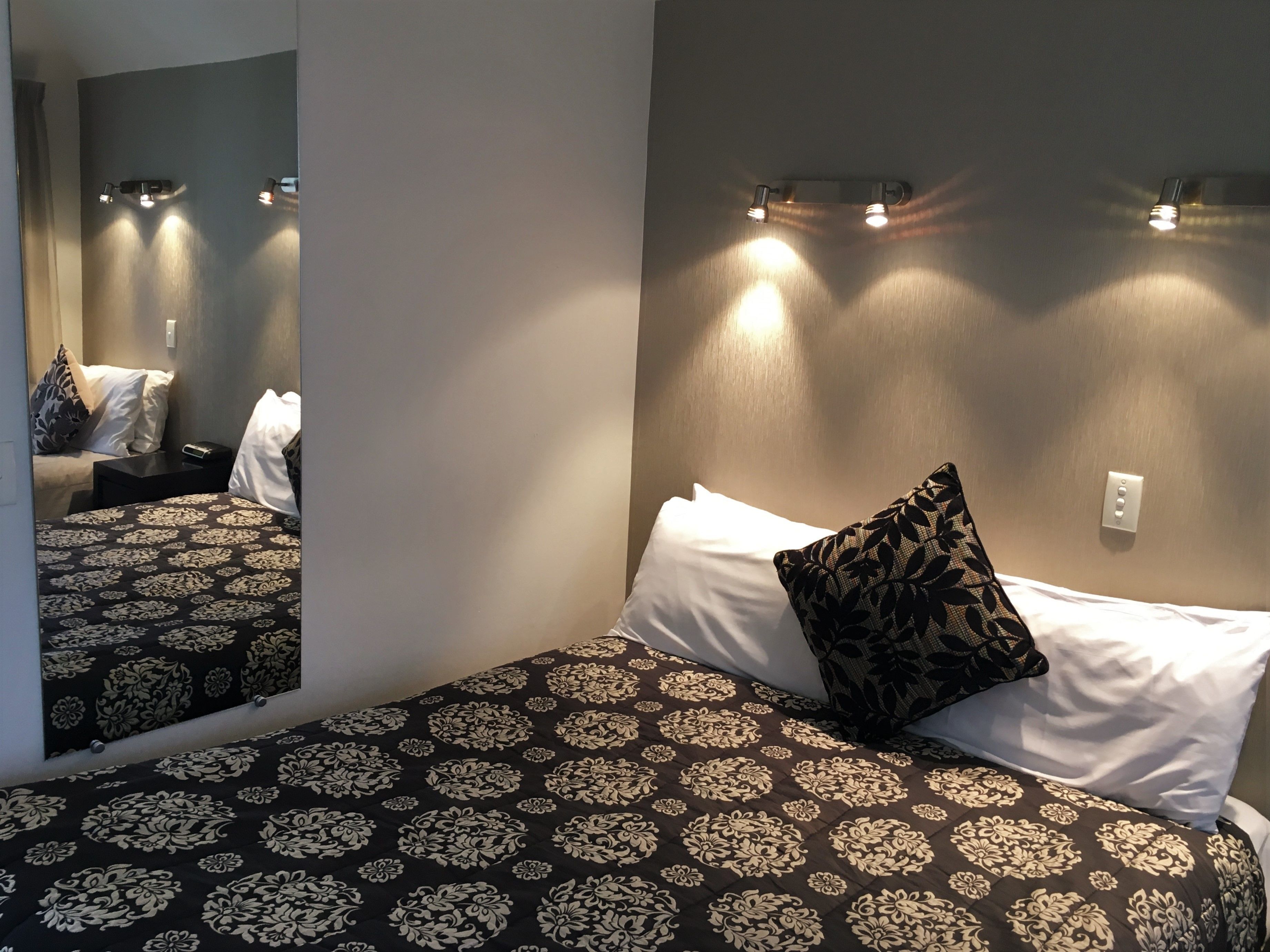 A hotel room with a bed, a nightstand, a mirror, and wall-mounted lights.