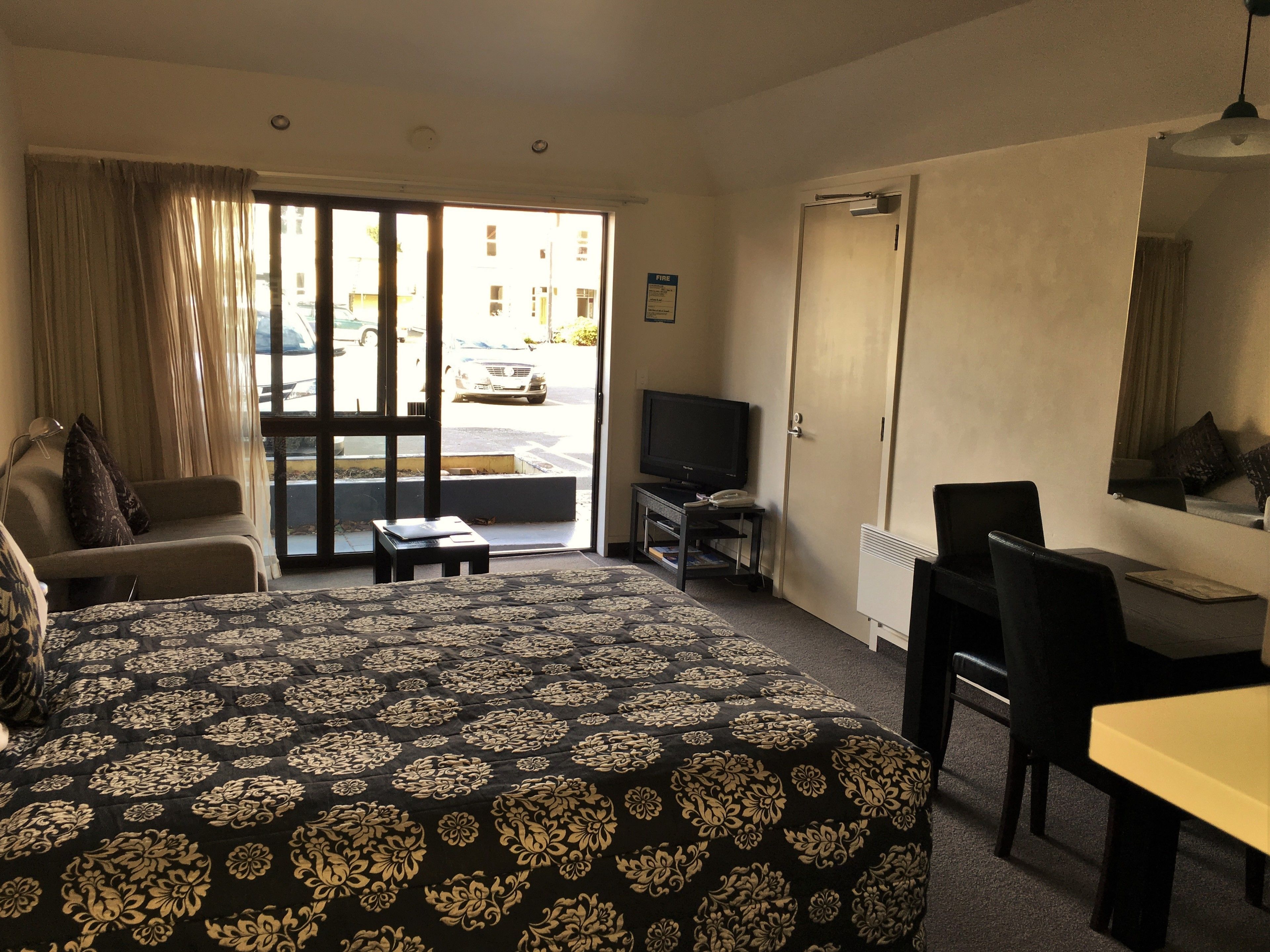 A hotel room with a bed, a desk, a chair, a television, and a view of the street outside.