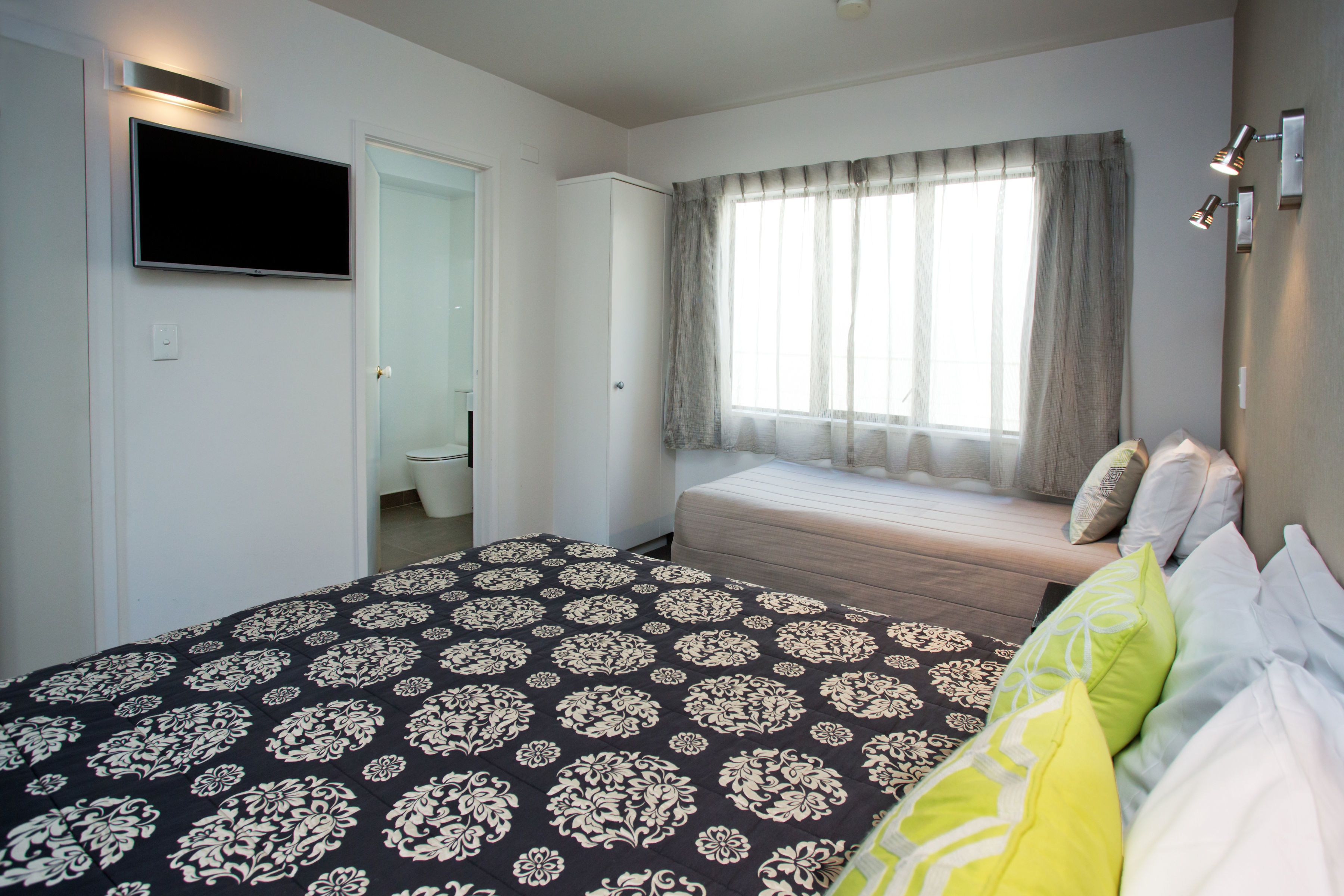A bedroom with a bed, a TV mounted on the wall, a window with curtains, and a bedside table with a lamp.