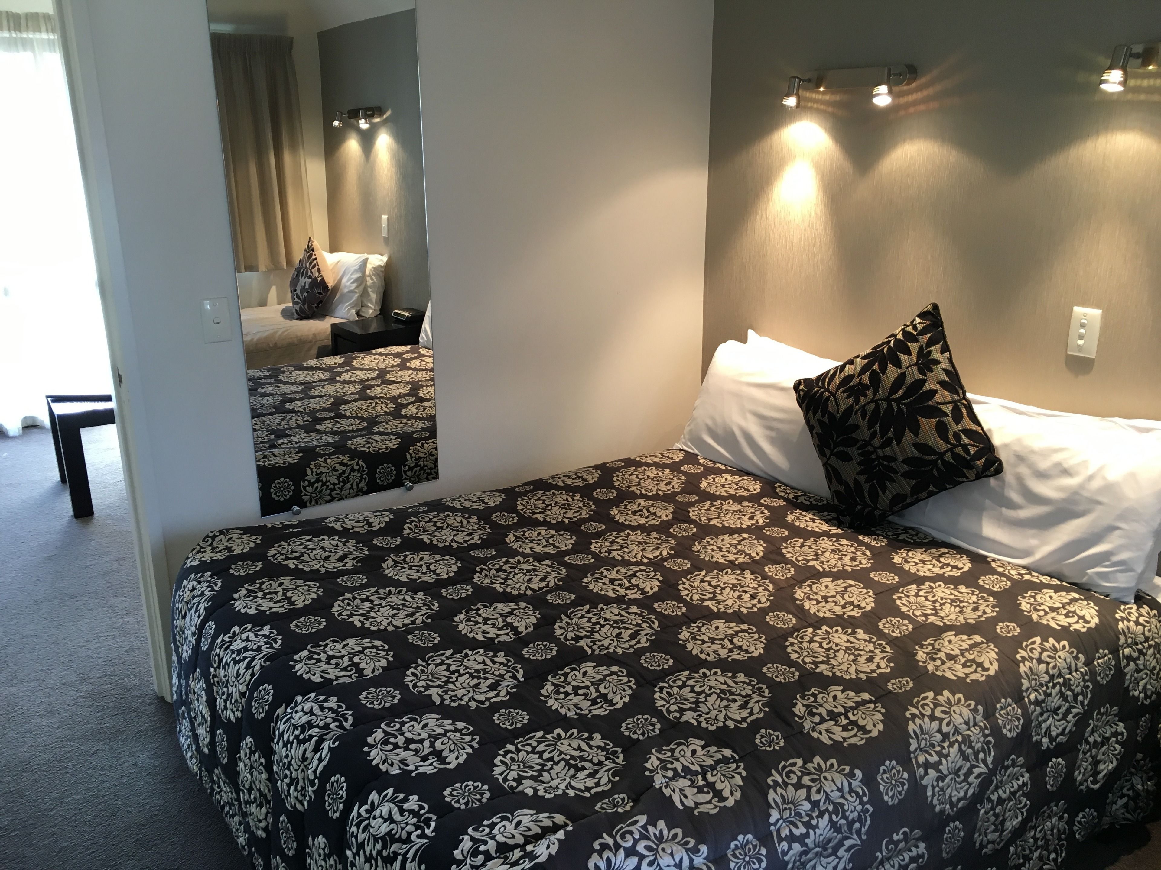 Una cama bien hecha con una colcha floral blanco y negro, un almohadón blanco y un almohadón estampado. Hay una mesita de noche con una lámpara y un espejo en la pared.