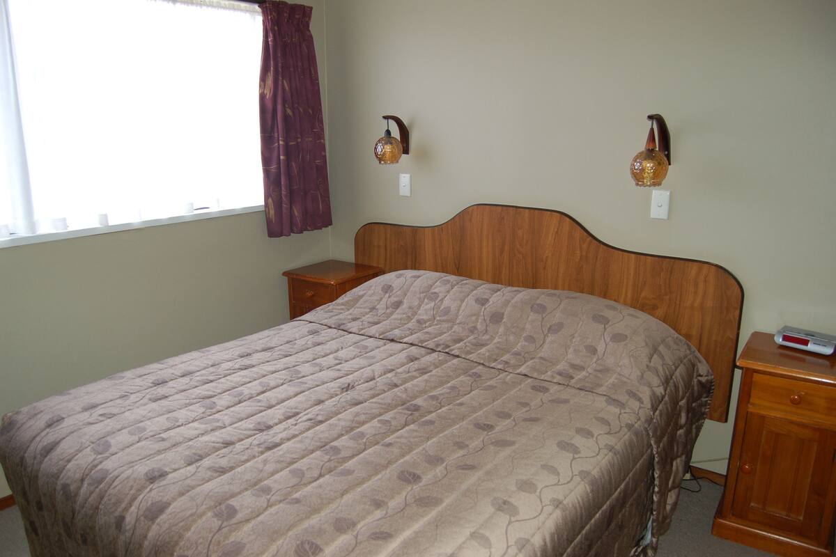 Un dormitorio con cabecera de madera, una cama con colcha estampada, dos mesitas de noche y dos lámparas de pared.