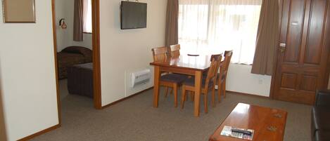A living room with a wooden dining table, four chairs, a television on the wall, and a door.