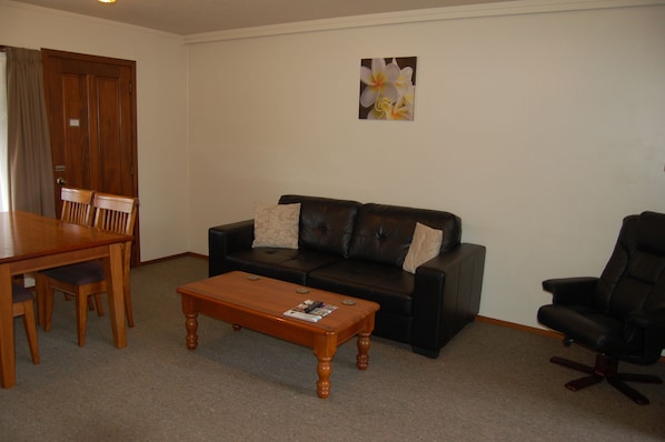 A living room with a wooden dining table, two chairs, a black leather sofa with cushions, a wooden coffee table, and a black office chair.