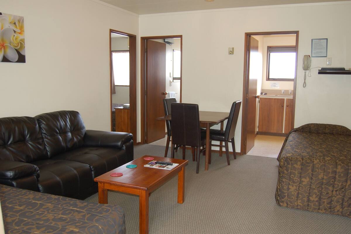 Sala de estar con sofá de cuero, mesa de centro de madera y sillón estampado. Hay un comedor con mesa y sillas, y una cocina con lavamanos y encimera a la vista.