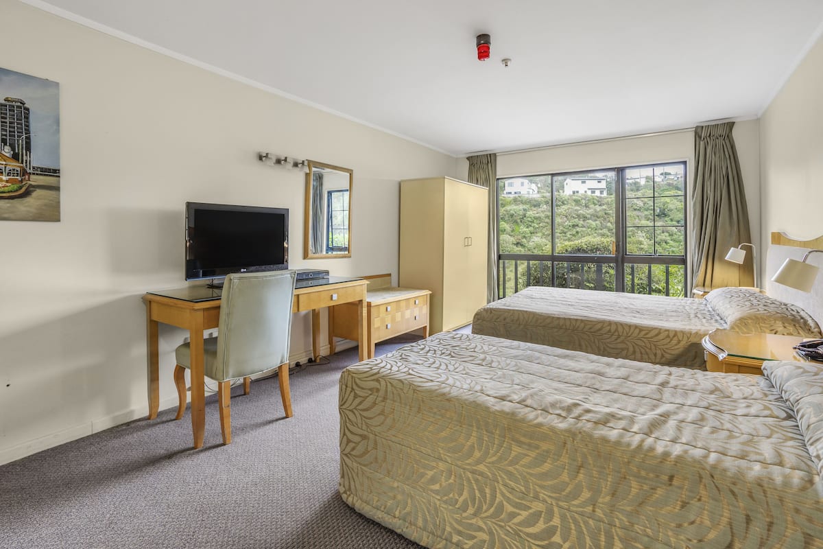 A hotel room with two beds, a desk with a computer, a chair, a wardrobe, and a large window with a view of greenery.
