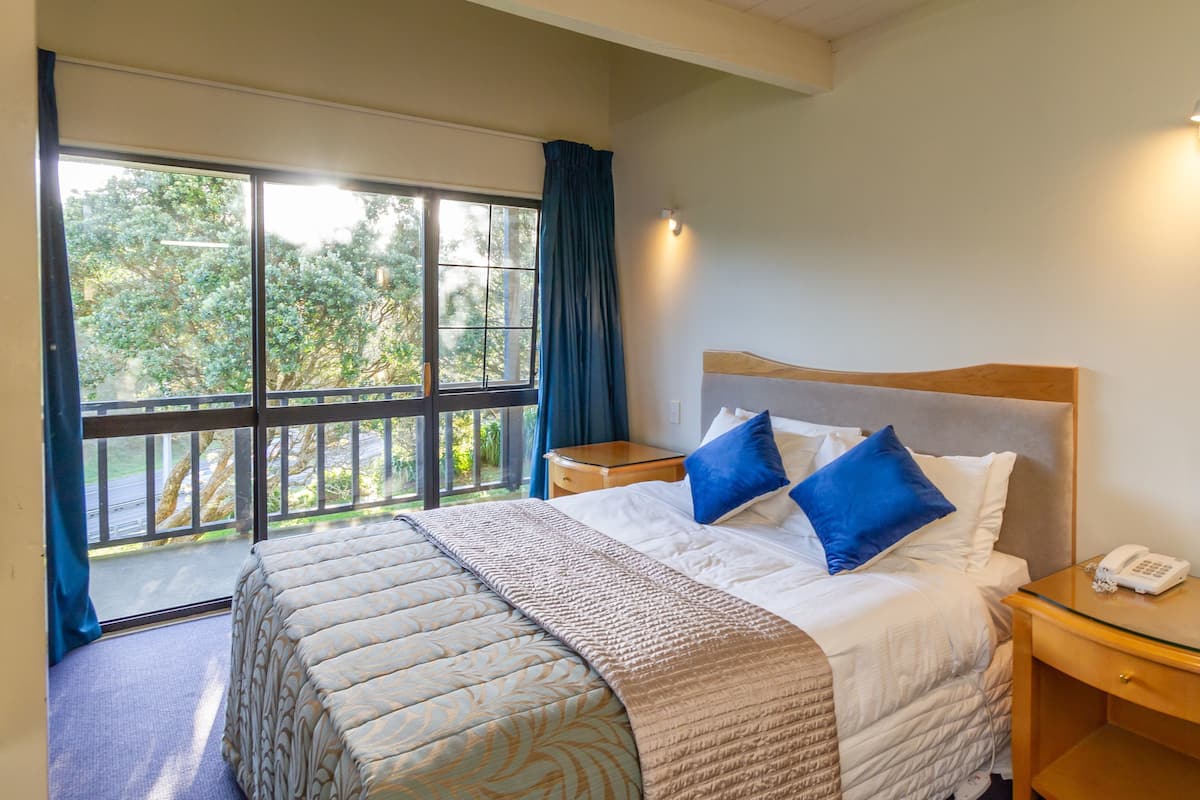 A hotel room with a large bed, a bedside table with a phone, and a view of greenery through a sliding glass door.
