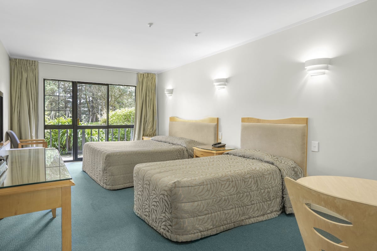 A hotel room with two beds, a glass table, a chair, a nightstand, and a view of greenery outside.
