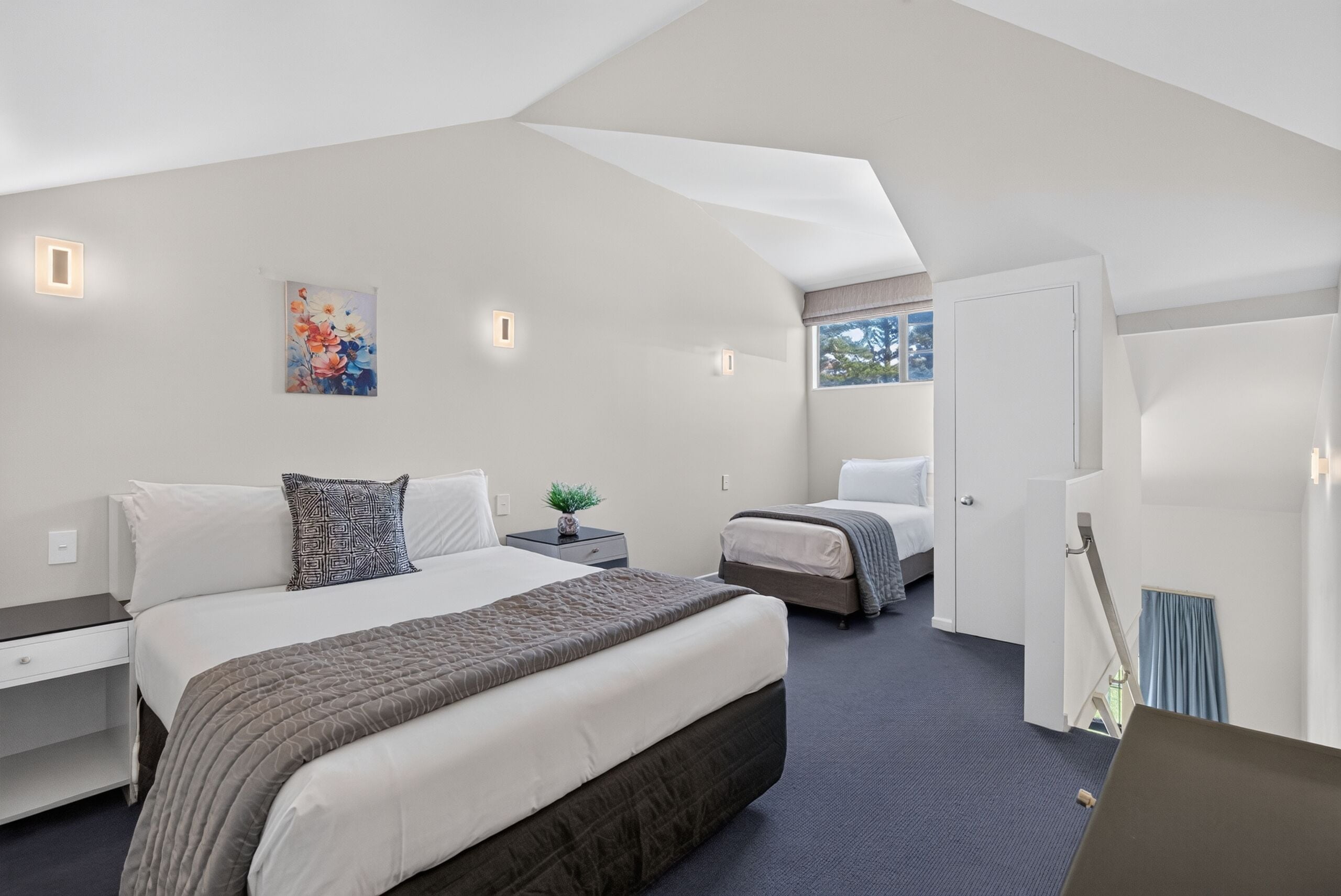 A modern bedroom with a large bed, a smaller bed, a nightstand with a plant, and a painting on the wall.