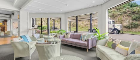 A spacious indoor area with a sofa, chairs, and a round table, featuring large windows and a view of the outside.