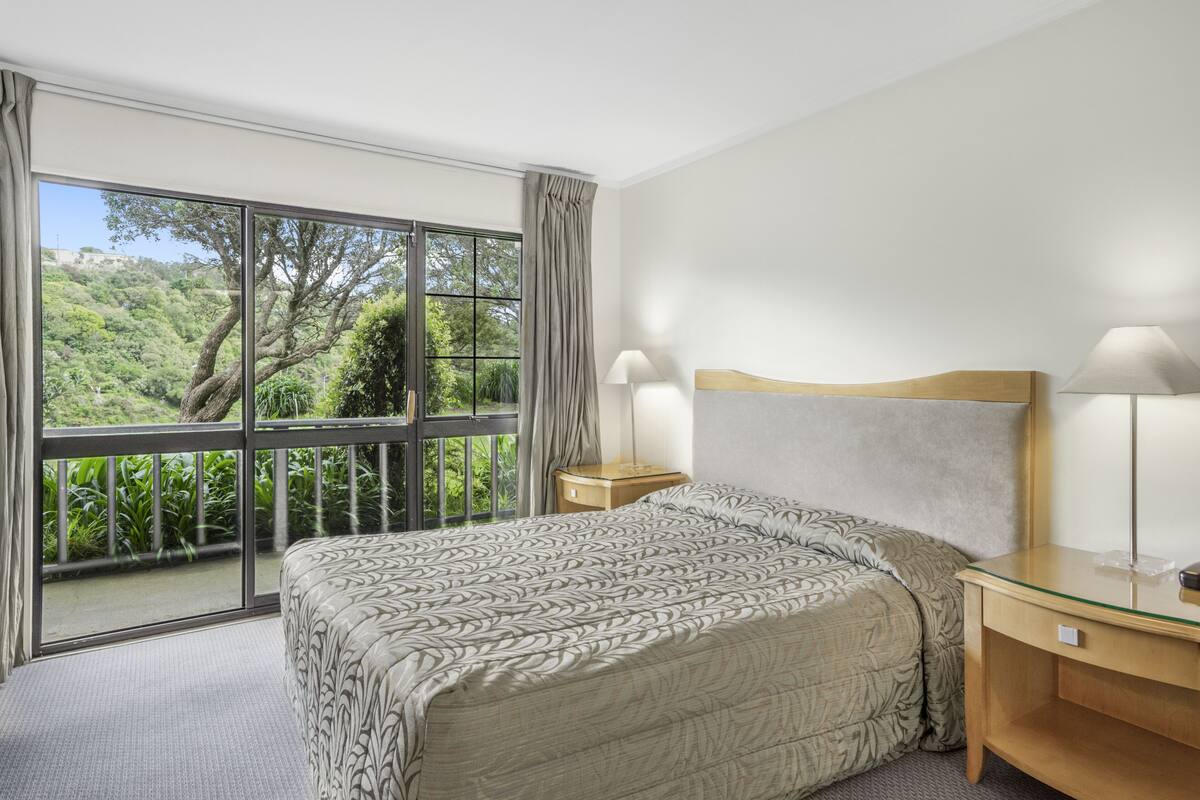 A bedroom with a large window, a bed with a patterned bedspread, two bedside tables with lamps, and a view of greenery outside.