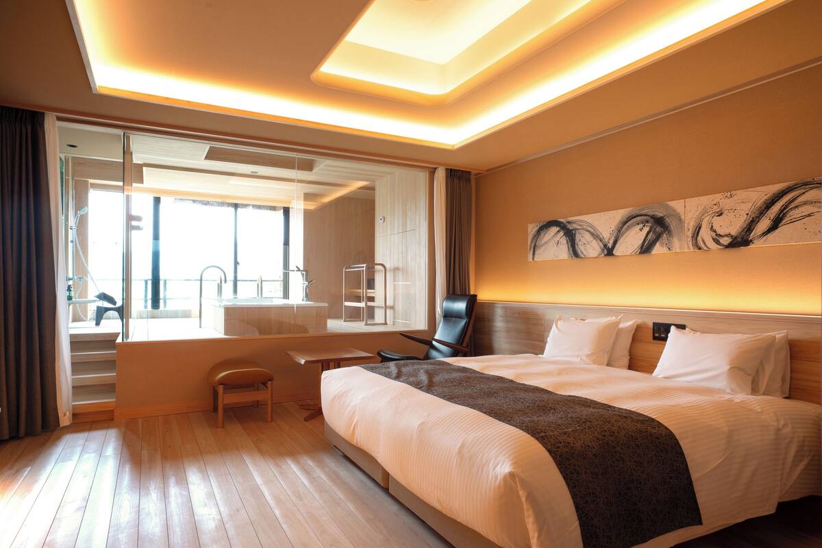 A modern hotel room with a large bed, a desk, and a chair. There is a view of a balcony and a bathroom with a mirror and a towel rack.