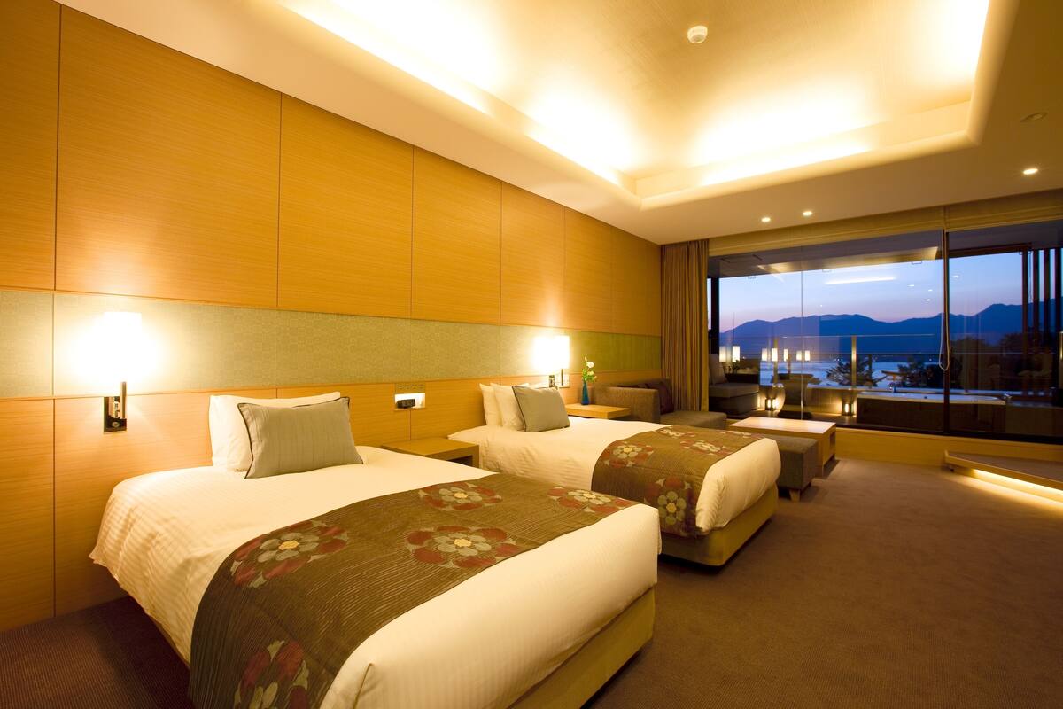 A hotel room with two beds, a large window with a view, and a balcony with a table and chairs.