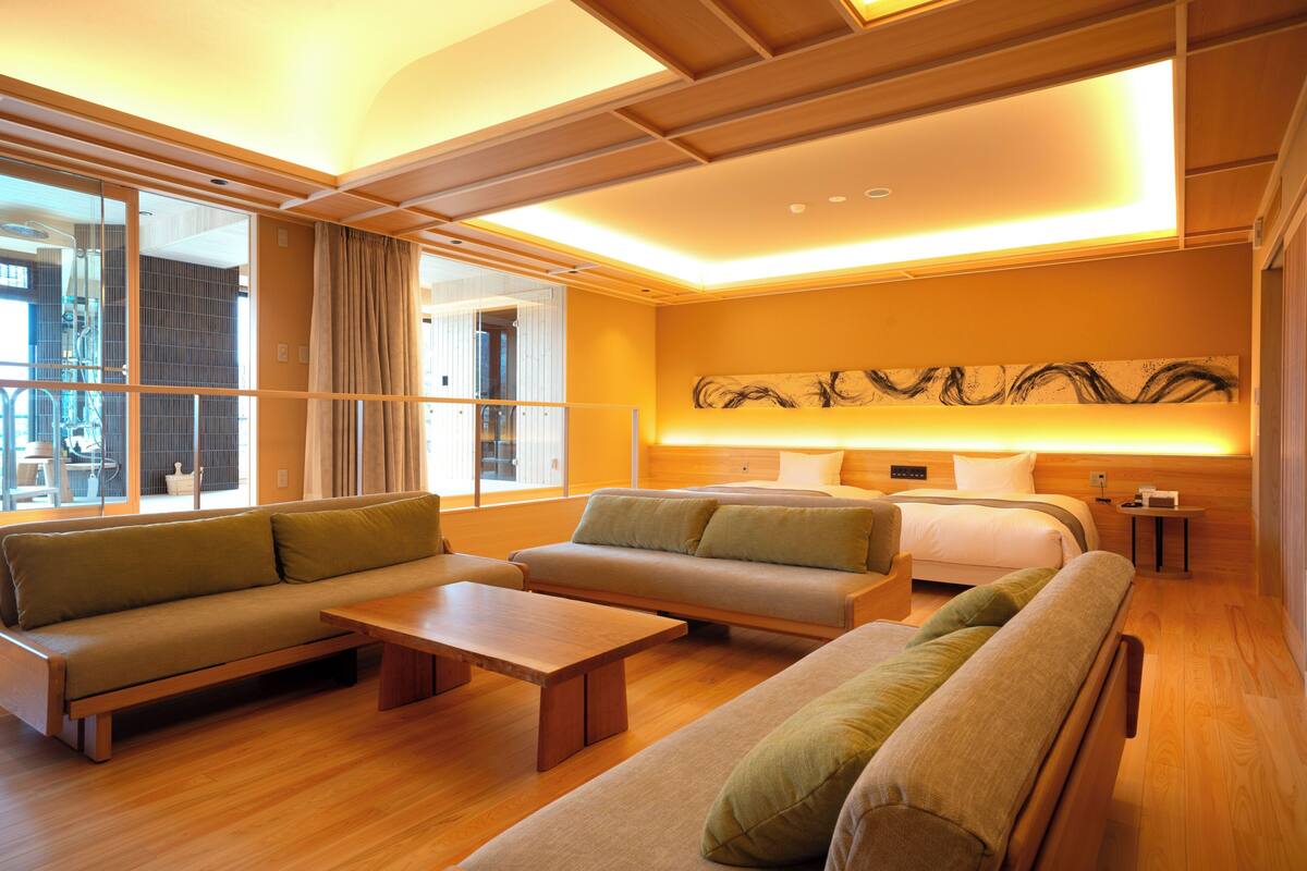 A modern hotel room with a sofa, a bed, a wooden coffee table, and a large window with curtains.