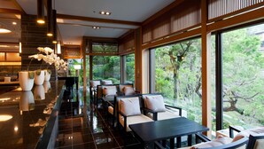 A modern hotel lobby with a long reception desk, seating area with cushioned chairs, large windows offering a view of greenery, and a polished floor. - Miyajima Grand Hotel Arimoto (Hatsukaichi)