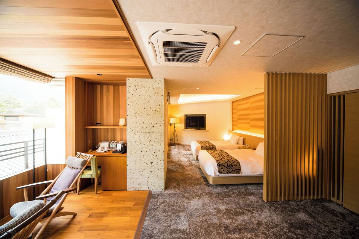 A hotel room with a wooden floor, a bed with a patterned blanket, a desk with a chair, a window, and a wall-mounted TV.
