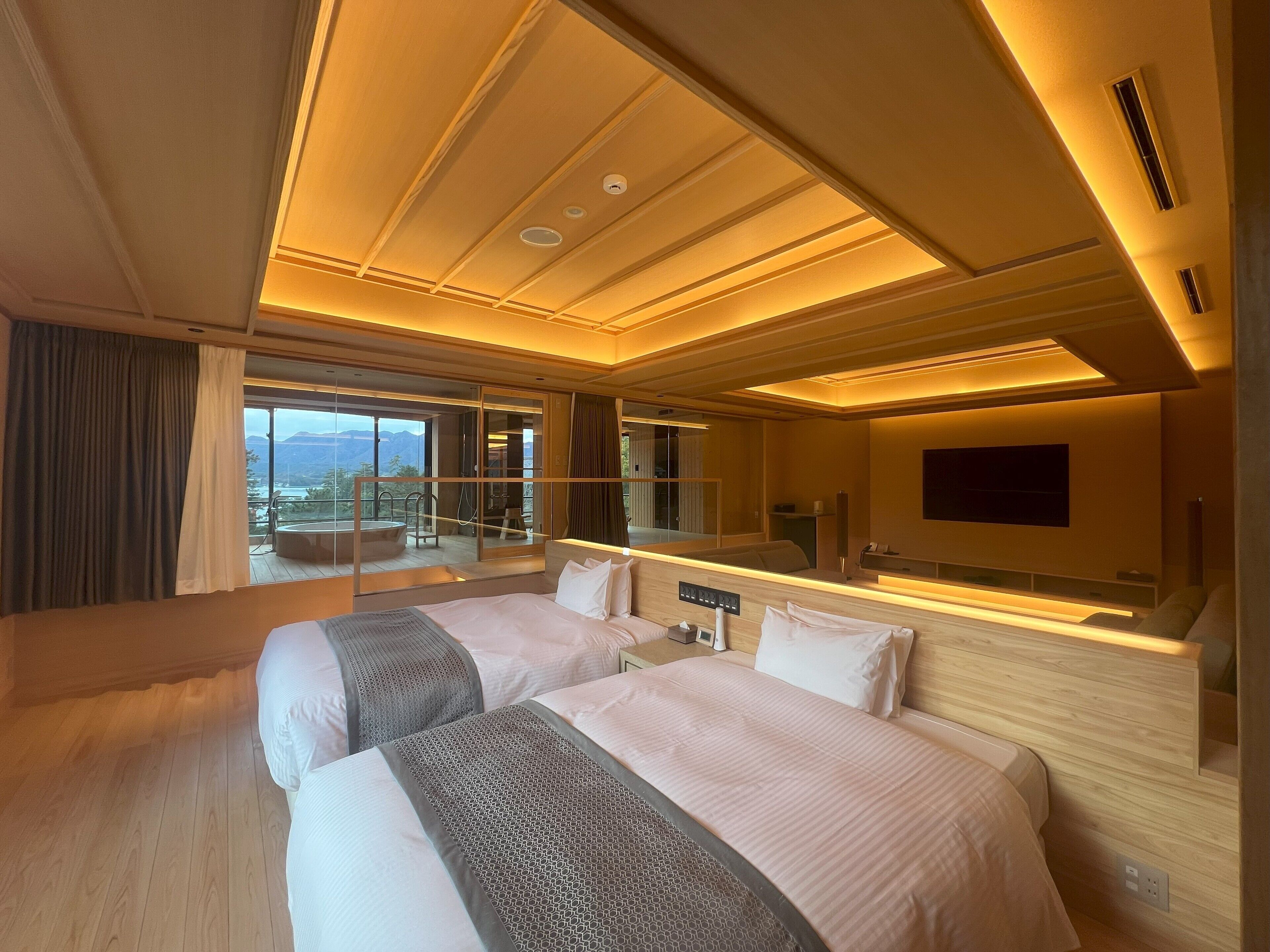 A hotel room with two beds, a large window with a view, a flat-screen TV, and a balcony with a jacuzzi.