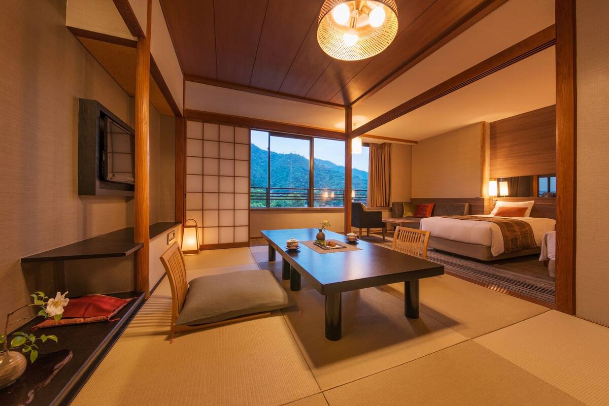 A traditional Japanese-style hotel room with tatami flooring, a low table with cushions, a bed with a view of the mountains, and a sliding door.