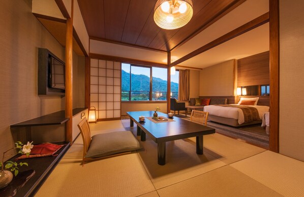 Habitación de hotel al estilo japonés tradicional, con suelo de tatami, una mesa baja con cojines, una cama con vistas a las montañas y una puerta corredera.