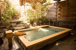 A hot spring bath area with stone steps leading into the water, surrounded by greenery and a wooden wall.