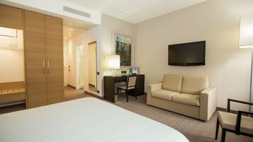 A hotel room with a large bed, a desk with a chair, a sofa, a TV mounted on the wall, and a lamp.