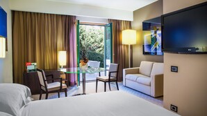 Quarto de hotel com cama, mesa de vidro, duas cadeiras, sofá, televisão e vista para o exterior.