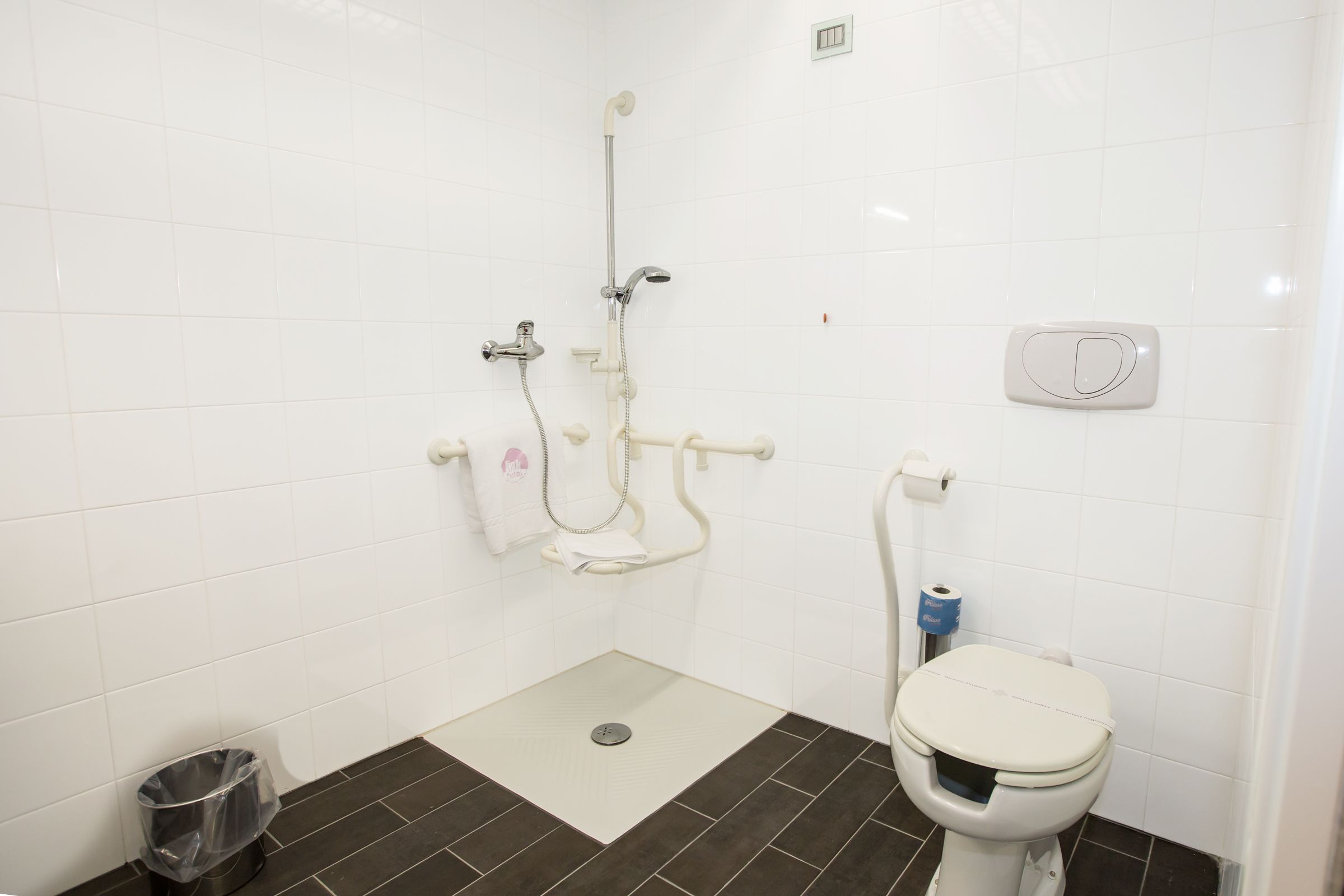 A bathroom with white tiled walls, a white toilet, a shower with a handheld showerhead, and a trash bin.