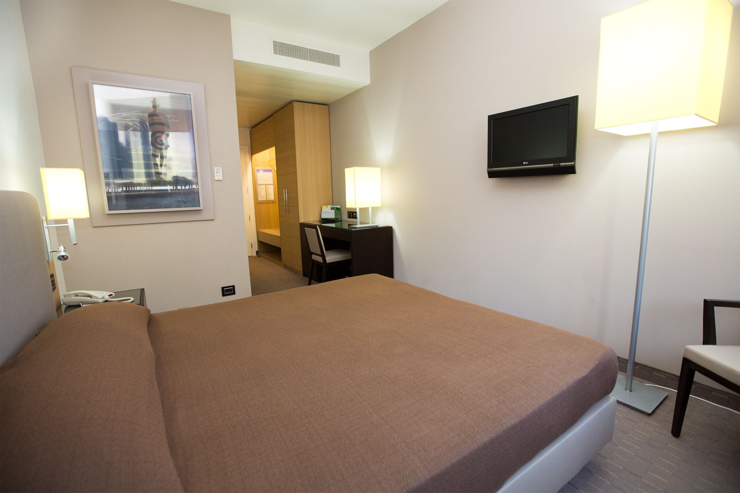 A hotel room with a bed, desk, chair, TV, lamps, and a framed picture on the wall.