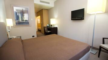 Habitación de hotel con cama, escritorio, silla, televisor, lámparas y un cuadro en la pared.