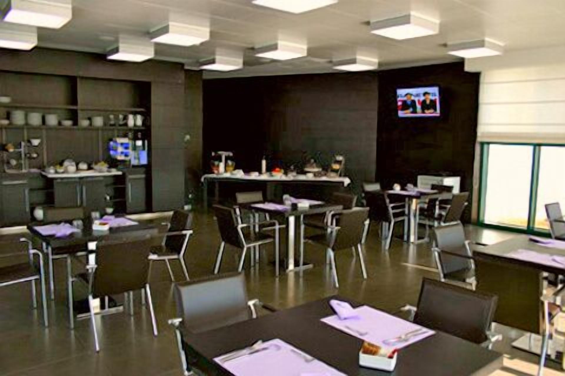 A conference room with tables and chairs, a television screen, and a kitchen area in the background.