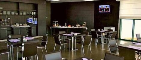A conference room with tables and chairs, a television screen, and a kitchen area in the background.