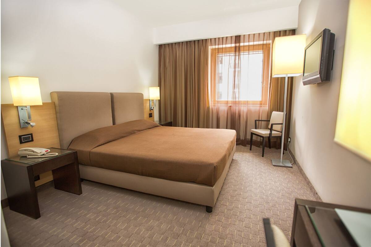 A hotel room with a large bed, two bedside lamps, a desk with a telephone, a chair, and a TV mounted on the wall.