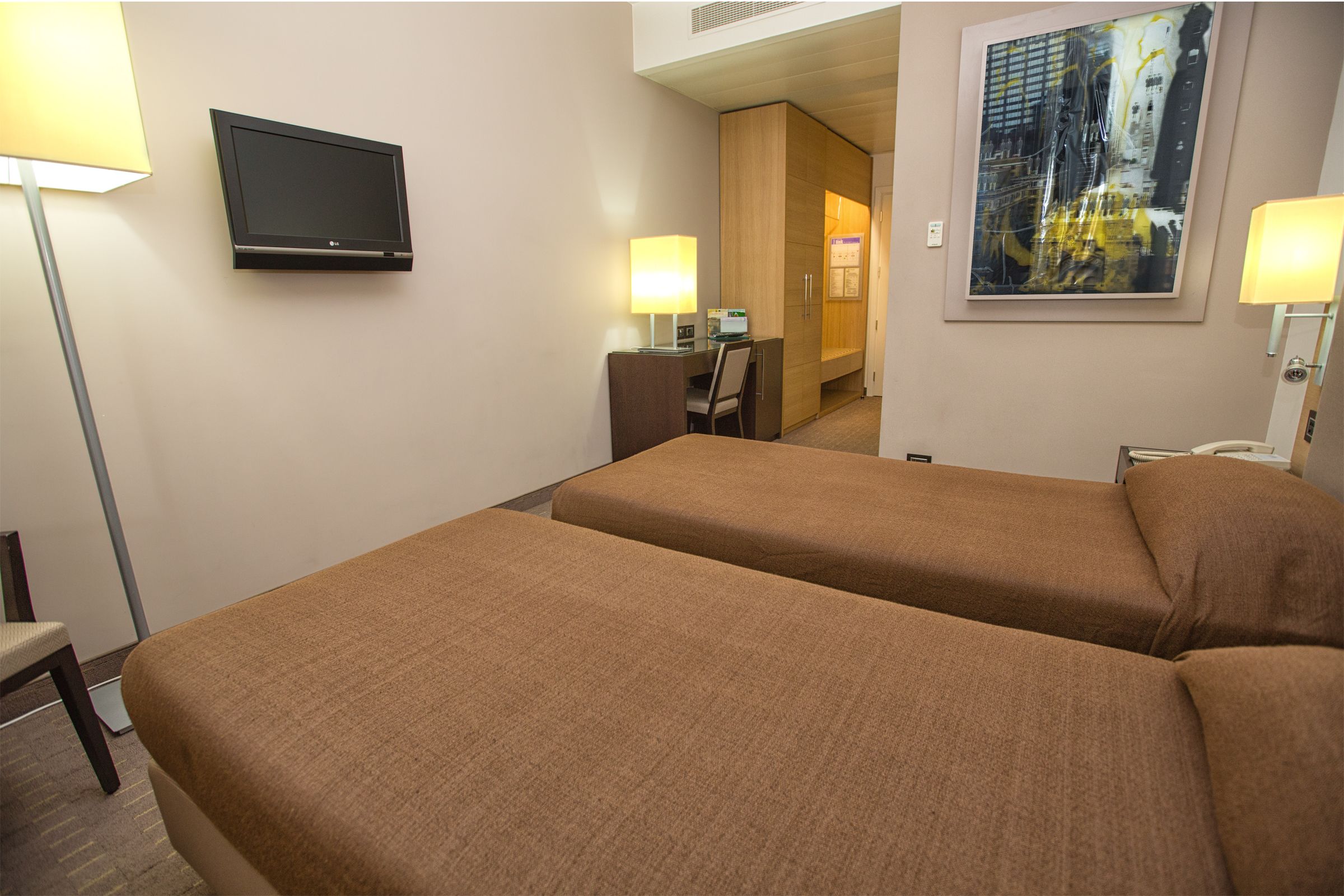 Ein Hotelzimmer mit zwei Betten, einem Fernseher, einem Schreibtisch mit Stuhl und einem Bild an der Wand.