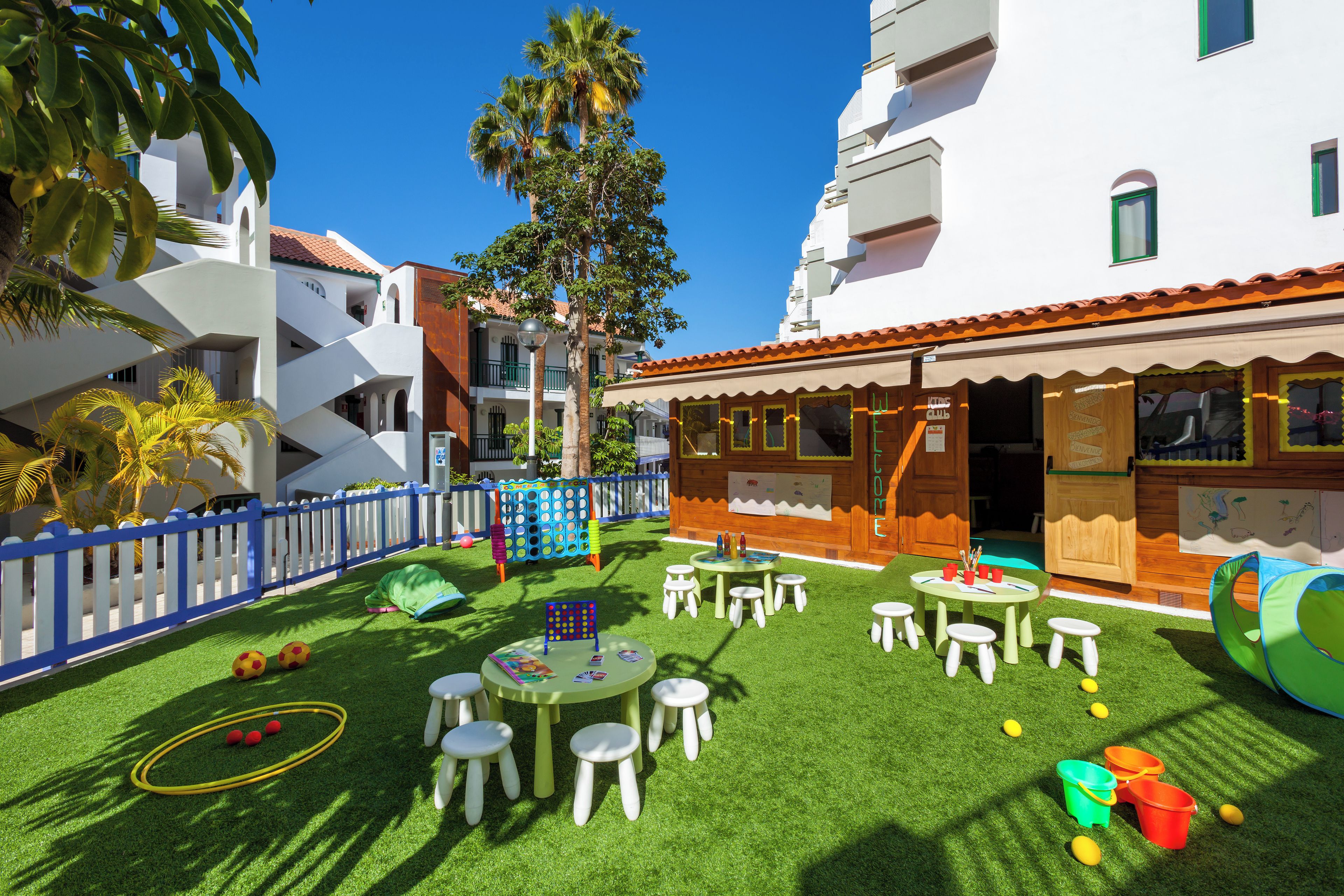 A children's play area with artificial grass, tables, and chairs, surrounded by a white fence and palm trees.