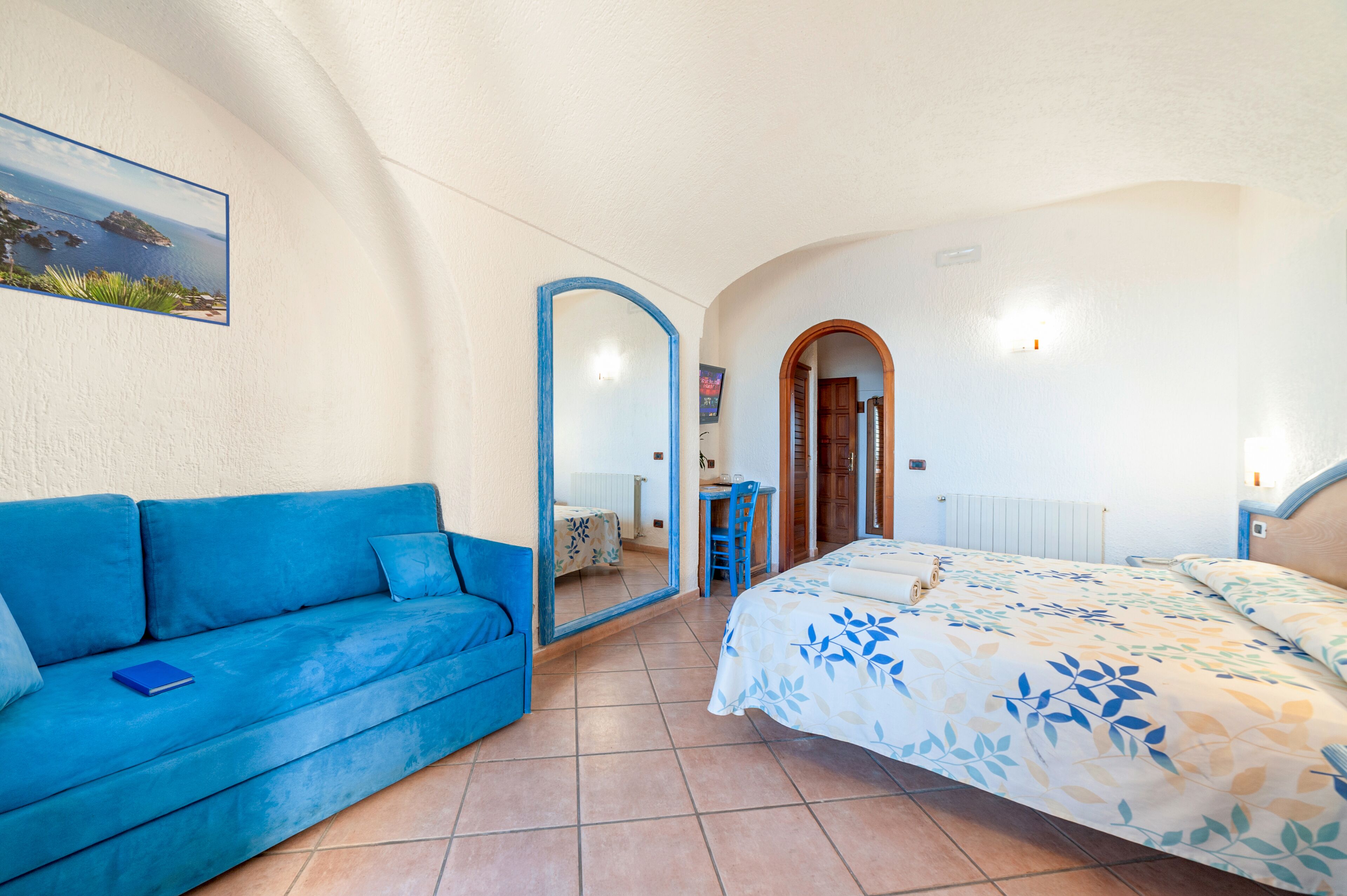 Un dormitorio con un sofá azul, una cama con colcha estampada, un espejo grande y vistas al mar a través de una ventana.