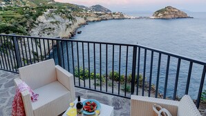 A balcony with wicker furniture, a table with food and drinks, and a scenic coastal view. - Punta Chiarito Resort (Forio)