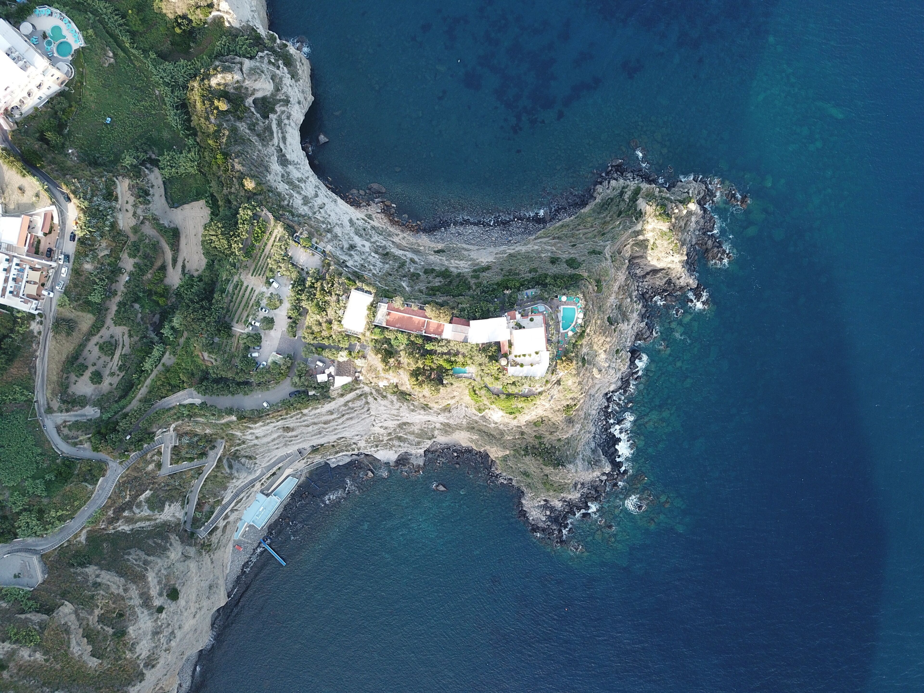 Vue aérienne d’une propriété située sur la côte, avec une route sinueuse, une piscine et un bâtiment doté d’un toit rouge.