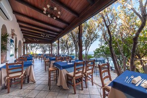 An outdoor dining area with tables and chairs, covered by a wooden roof, overlooking a scenic view.